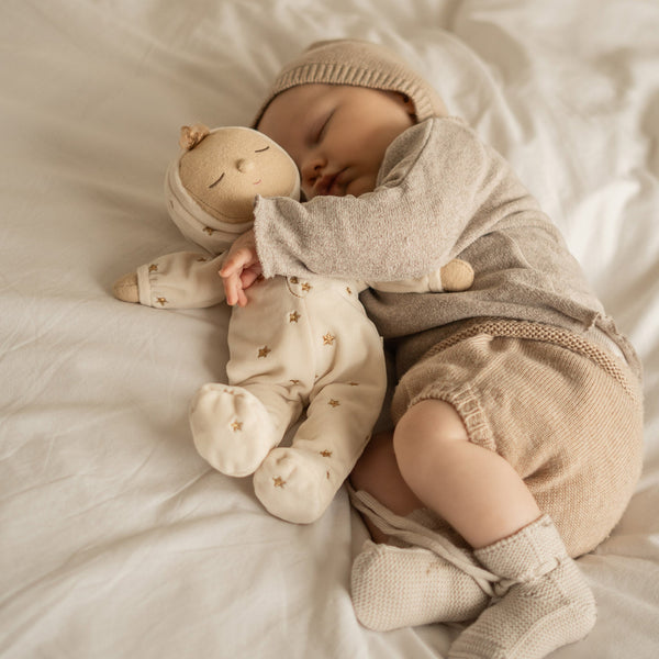 Lullaby Dozy Dinkum - Lyra soft doll cuddled by a sleeping child on a bed in a cosy neutral outfit with a bonnet and star print pajamas from Dozy Dinkums