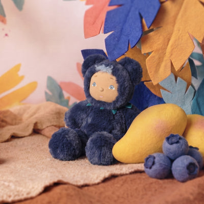 Cozy Dinkum - Blueberry Bear by Dinkum Dolls by Olli Ella sitting beside blueberries and a yellow pear in a playful jungle scene