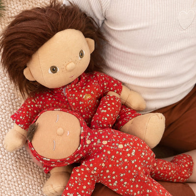 Two Dinkum Dolls in matching red floral outfits held on a child’s lap: a freckled doll with auburn hair cradles a sleeping baby doll.