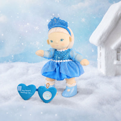 Plush doll in a blue sequinned dress and tiara stands in snow beside a blue heart accessory and a snowy cottage.