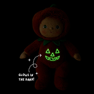 A plush doll dressed in a red pumpkin costume with a glowing green jack-o'-lantern face on the front, shown on a black background with the text 'GLOWS IN THE DARK!' and a starburst graphic.