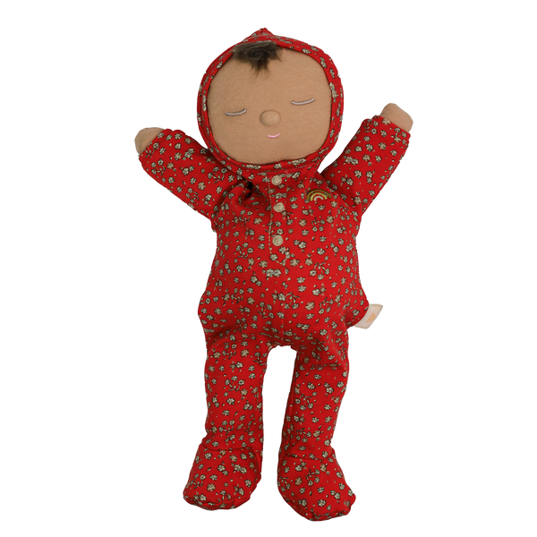 Soft baby doll with sleepy embroidered closed eyes, a smiling mouth and a tuft of dark brown hair, wearing a red footed sleepsuit and matching bonnet with a ditsy floral print, button placket, a small embroidered rainbow, and arms raised.