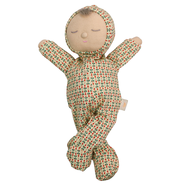Soft baby doll with sleepy embroidered closed eyes, a smiling mouth and a tuft of brown hair, wearing a cream footed sleepsuit and matching bonnet printed with small red and green sprigs, with arms raised and legs crossed.