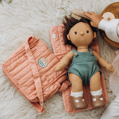 Carrie Convertible Changing Set - Rose by Dinkum Dolls with a soft doll in a green romper on a quilted changing mat beside a matching carry bag and brush