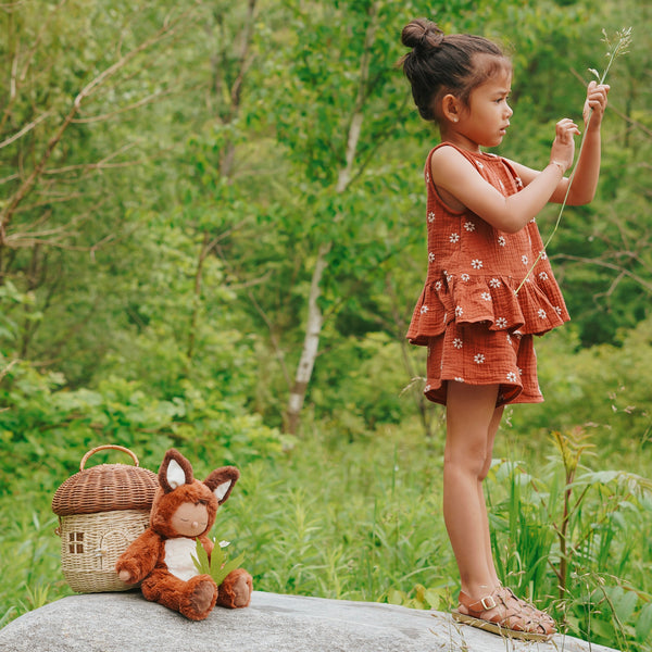 Cozy Dinkums - Fox - Finnley fox plush doll sitting beside a woven basket outdoors while a child explores nature