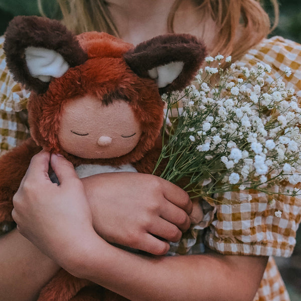 Cozy Dinkums - Fox - Finnley rust-colored fox soft doll held by a child beside delicate white flowers from Dozy Dinkums