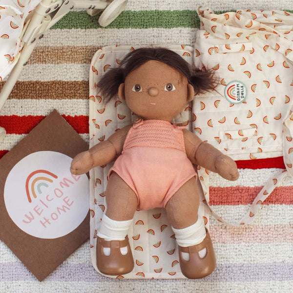 Dinkum Dolls - Dottie soft doll with brown hair wearing a peach romper white socks and brown shoes on a rainbow patterned play mat