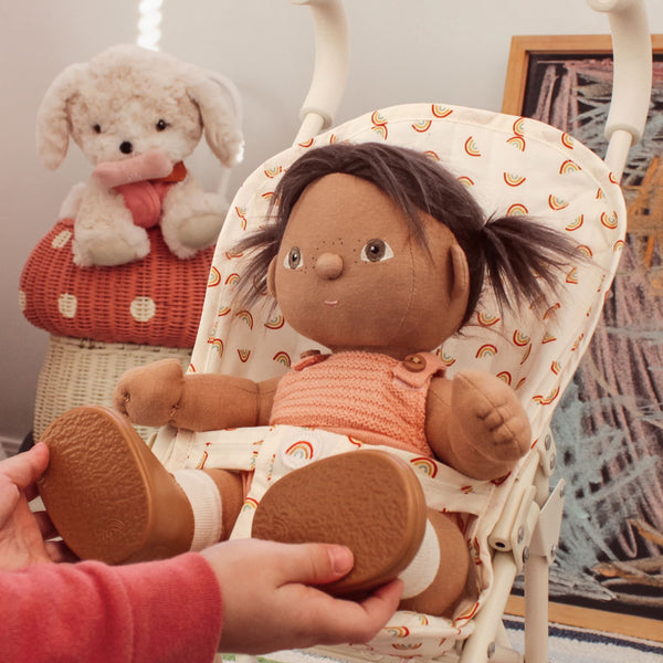 Dinkum Dolls - Dottie soft doll in a rainbow print pram with a child holding her feet and a plush dog nearby
