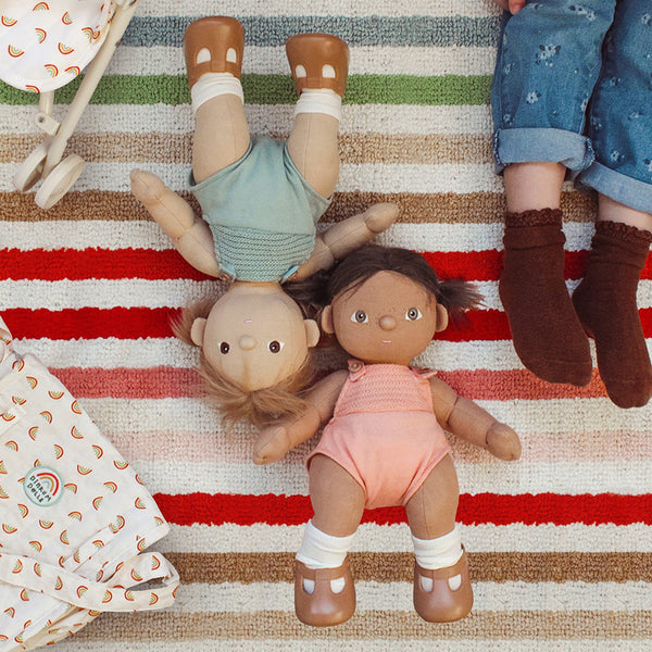 Dinkum Dolls - Dottie soft doll with brown hair wearing a pink outfit and white socks lying beside other dolls on a striped rug for imaginative play