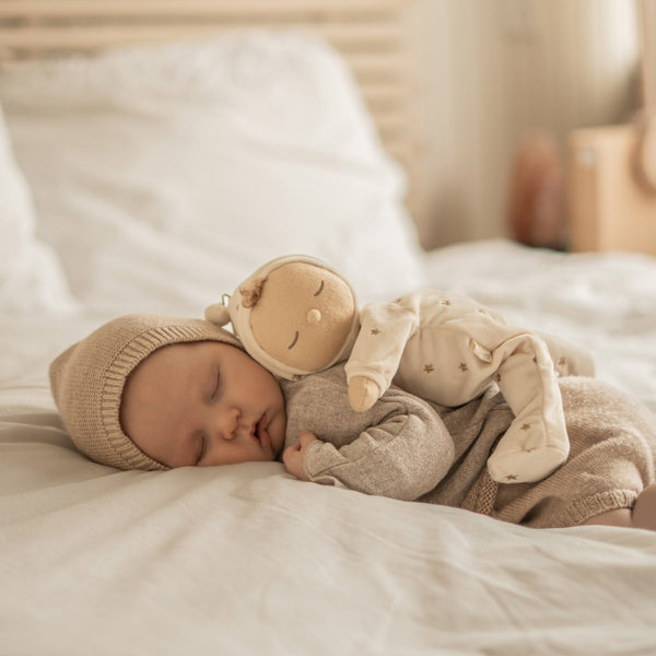 Lullaby Dozy Dinkum - Lyra soft doll cuddled by a sleeping child in bed wearing a bonnet and neutral outfit from Dozy Dinkums for gentle play