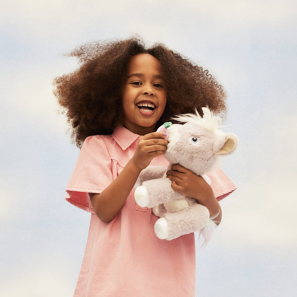 Dinkum Unicorn - Glimmer by Dinkum Unicorns cuddly lilac plush unicorn held by a smiling child in a pink dress