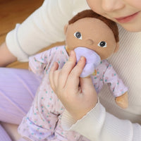 Child holding a plush toy with a pacifier, wearing a white shirt and purple pants.