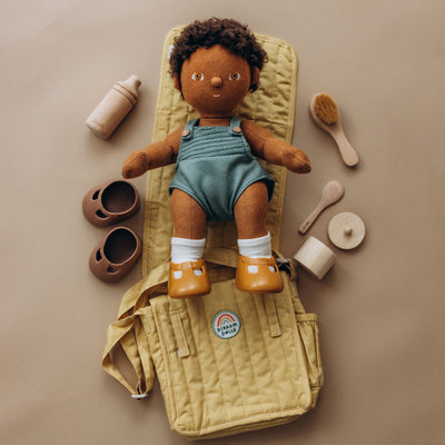 Carrie Convertible Changing Set - Honey by Dinkum Dolls with a soft doll on a quilted changing mat surrounded by a bottle shoes brush and care accessories