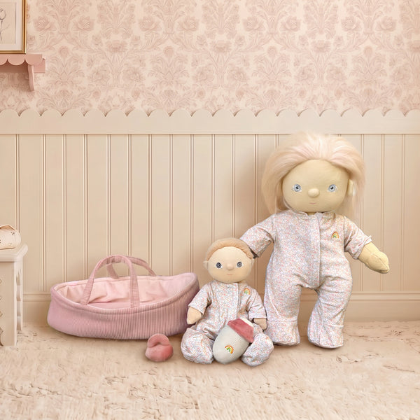 Two dolls in matching outfits standing next to a pink bassinet in a nursery setting.