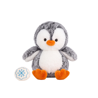 Fluffy plush penguin with grey speckled fur, a white face and tummy, black eyes, and an orange beak and feet, sitting beside a small cream fabric snowball embroidered with a blue snowflake.