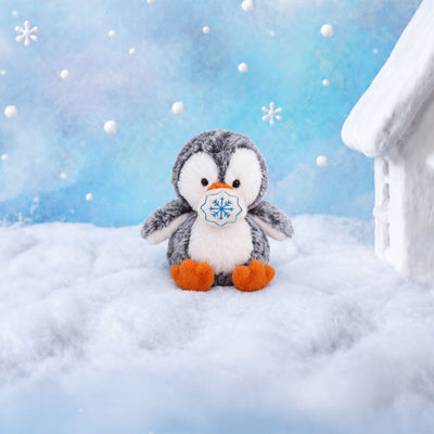 Fluffy plush penguin with grey speckled fur, a white face and tummy, black eyes, and an orange beak and feet, sitting beside a small cream fabric snowball embroidered with a blue snowflake.