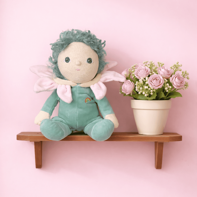 Stuffed toy with green hair and outfit on a wooden shelf against a pink wall with a potted plant.