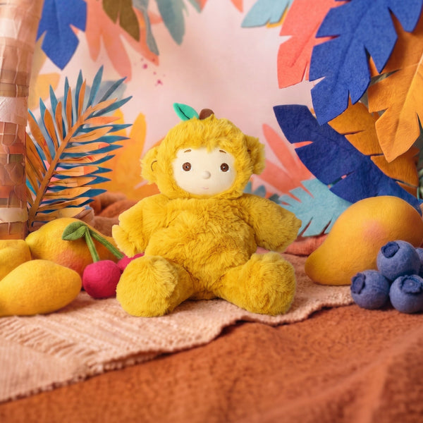 A yellow plush monkey toy with a white face sits on a textured blanket surrounded by colorful tropical paper leaves and fruit props against a patterned backdrop.