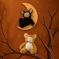Two plush animal toys styled in a decorative scene: a black cat-like plush sitting on a gold crescent moon and an orange cat-like plush sitting on a brown branch, against a warm brown background with small gold stars.