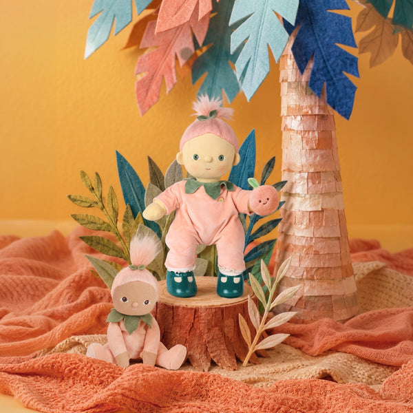 A styled product display featuring two plush dolls in pink outfits, one standing on a wooden stump and one sitting on a peach-colored blanket, with a paper palm tree and tropical leaves in the background against a warm yellow wall.