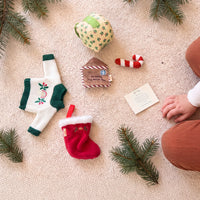 Dinkum Doll Christmas accessories laid on a cream knit rug with pine sprigs: a knitted holly jumper, red stocking, plush gift box, candy cane, letter to Santa and a note card.