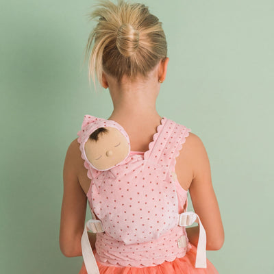 A young girl viewed from behind wearing a pink polka-dot baby carrier wrap with a matching bonnet featuring a sleeping doll face, set against a solid pale green background.