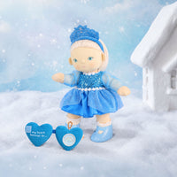 Plush doll in a blue sequinned dress and tiara stands in snow beside a blue heart accessory and a snowy cottage.
