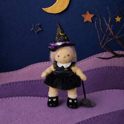 A small witch doll with a black dress, pointed witch hat, and black shoes stands on a purple hill against a dark blue paper-cut sky with a gold crescent moon, stars, and bare branches.