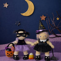 Two Halloween-themed dolls posed on a purple textured surface against a dark blue backdrop with a gold crescent moon, stars, and a bare tree branch. One doll wears a black costume with a purple skirt and holds an orange jack-o'-lantern bucket.