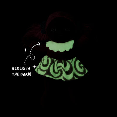 A dark, glow-in-the-dark doll or plush toy shown against a black background, with a bright neon green ruffled collar and a glowing green and black patterned dress. White text on the left reads 'GLOWS IN THE DARK!' with an arrow pointing to the outfit.