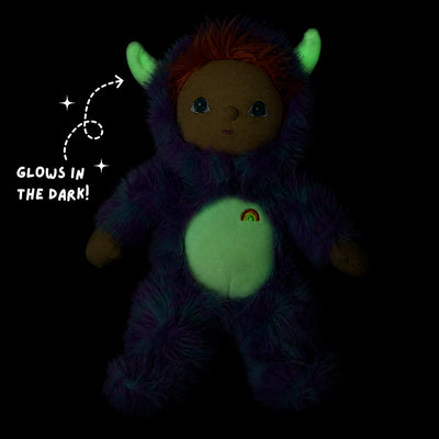 A plush monster doll with glowing green belly and horns, dark purple fur, and a small rainbow emblem on the chest, shown on a black background with the text 'Glows in the dark!'.
