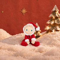 A small plush doll dressed in a red Santa-style outfit with white trim and a red hat sits on a cream textured surface against a warm red-orange backdrop, with decorative gold stars and lit Christmas trees behind it.