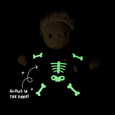 A dark plush doll with short blonde hair wearing a black outfit that glows green with a skeleton bone and rib pattern, shown on a black background with the text 'GLOWS IN THE DARK!'.