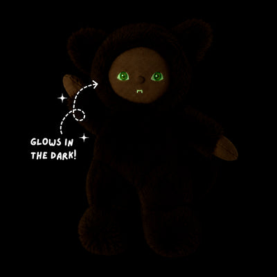 A dark plush teddy bear with glowing green eyes and a smaall stitched smile, shown on a black background with the text 'GLOWS IN THE DARK!' and sparkles pointing to its eyes.