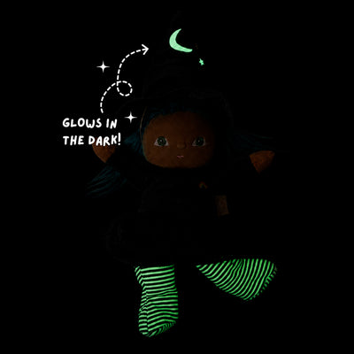 A dark plush doll wearing a black outfit and glowing green striped socks, with a glowing green crescent moon and stars on the hat. White text reads 'GLOWS IN THE DARK!' on a black background.