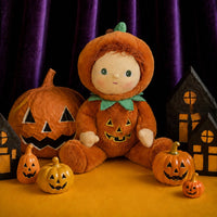 A plush Halloween doll dressed in an orange pumpkin costume with a green bow tie, seated on a yellow surface with carved jack-o'-lantern pumpkins and small house decorations against a dark purple curtain background.