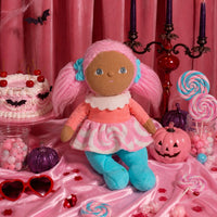 A plush doll with brown fabric skin, pink yarn hair styled in pigtails with blue bows, wearing a pink and white patterned dress and blue boot covers