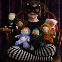A child wearing a black cat-eye mask and black-and-white striped tights sits cross-legged on a wooden chair, holding several plush dolls against a dark purple velvet curtain background.