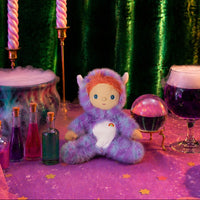 A plush fantasy doll with orange hair, blue eyes, and a purple fluffy outfit sits on a pink starry tablecloth. The scene is styled with a green velvet curtain backdrop, pink-and-white twisted candles, small potion bottles, a glowing orb, and a purple wine glass.