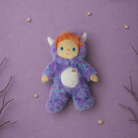 A plush toy dressed as a furry monster in a purple and blue onesie with orange hair, white horns, and a white belly patch, centered on a lavender background with decorative stars, clouds, and branches.