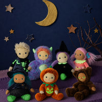 A group of seven plush Halloween dolls dressed in various costumes, arranged on a purple surface against a dark blue background decorated with a gold crescent moon and wooden stars.