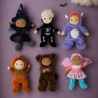 Six small plush dolls dressed in Halloween costumes arranged on a lavender background with decorative clouds and bats. The costumes include black witch, black skeleton, purple monster, orange pumpkin, brown bear, and pink fairy outfits.