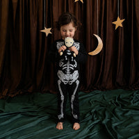 A young child stands barefoot on a dark green velvet floor in front of a brown curtain backdrop, wearing a black and white skeleton-print pajama set and holding a small plush toy. Decorative hanging stars and a crescent moon are visible behind the child.