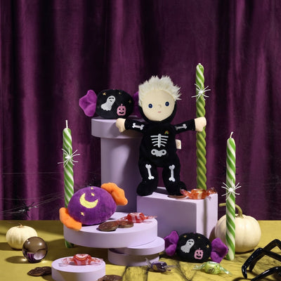 A Halloween-themed plush toy of a skeleton child in a black skeleton costume stands on a lavender pedestal, surrounded by candy, green-and-white striped candles, small pumpkins, and other spooky decorations against a dark purple curtain backdrop.
