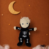 A Halloween plush doll dressed in a black skeleton jumpsuit with white bone details, posed against a textured brown background with a yellow crescent moon, stars, fake white cobwebs, and a small black spider decoration.