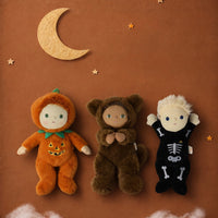 Three plush Halloween costumes displayed on a warm brown background with a paper crescent moon and stars. The left plush is orange with a jack-o'-lantern design and green collar.