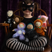 A child wearing a black cat-eye mask and black-and-white striped tights sits cross-legged on a wooden chair, holding several plush dolls against a dark purple velvet curtain background.