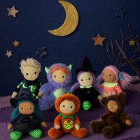 A group of seven plush Halloween dolls dressed in various costumes, arranged on a purple surface against a dark blue background decorated with a gold crescent moon and wooden stars.