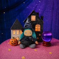 A Halloween-themed doll with blue hair wearing a witch hat and black dress sits between two miniature houses, with a carved pumpkin and a purple crystal ball on a pink surface against a starry purple backdrop.