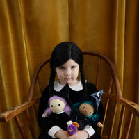 A young girl with black braided hair sits in a wooden chair against a gold velvet curtain, wearing a black dress with a white collar and holding two small dolls.